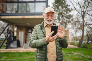 An elderly man with a beard stands in a yard in front of a house and smiles as he looks at his cell phone.